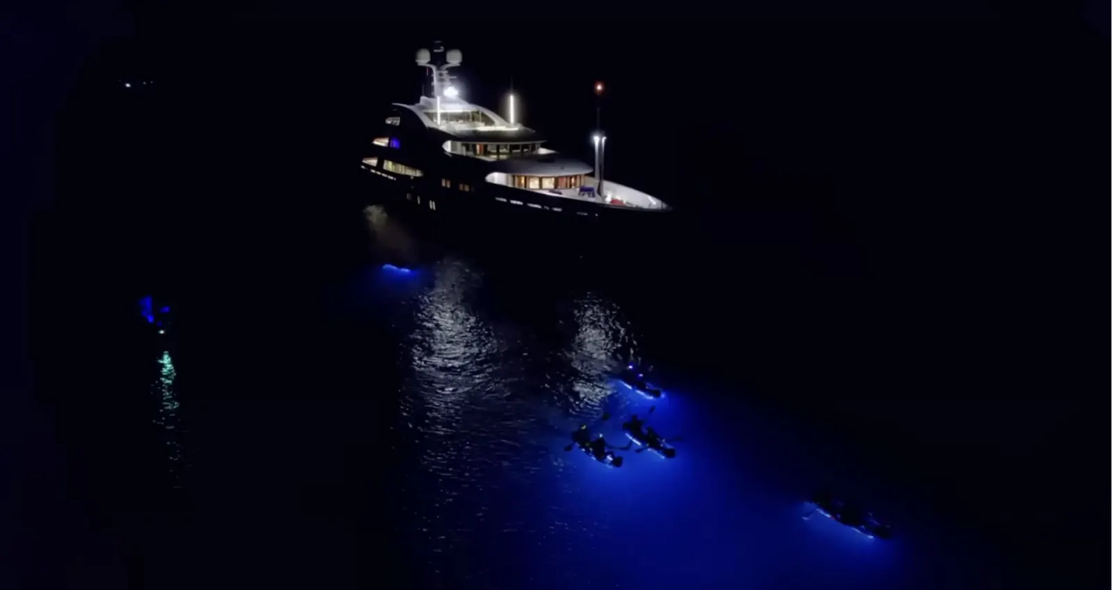 Illuminated clear kayaks rafted beside a brightly lit superyacht at night